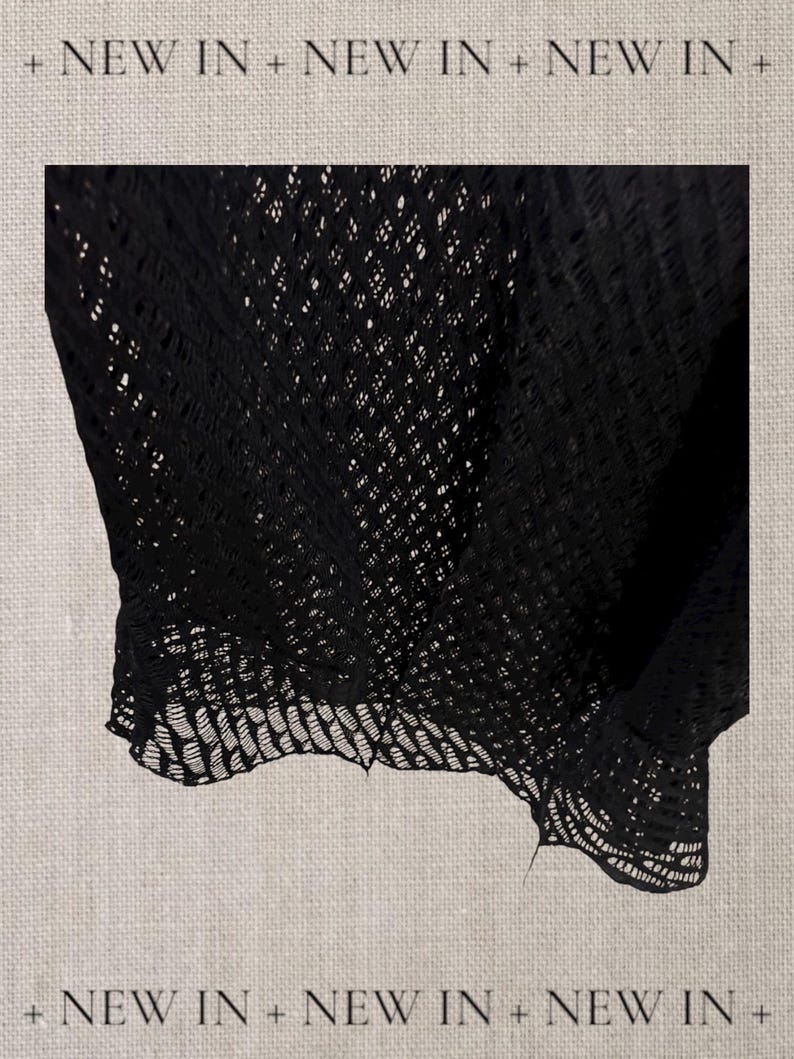 May include: Black open-weave fabric, possibly a top or shawl, with a delicate, lacy pattern. The fabric drapes softly, showcasing the intricate design. The words "NEW IN" are repeated across the top and bottom of the image.