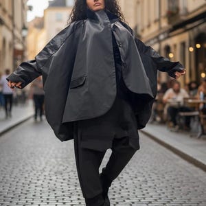 Pode incluir: Uma pessoa vestindo um traje preto, incluindo uma jaqueta tipo capa, calças e botas de plataforma, está em uma rua de paralelepípedos. A jaqueta tem um design único e grande com gola alta e fechamento por botões. O estilo geral é moderno e ousado.