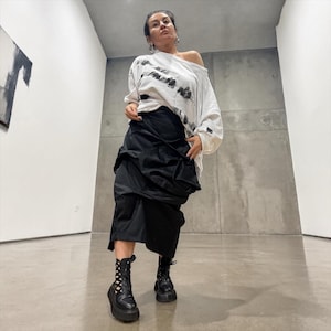 Könnte beinhalten: Eine Frau trägt ein weißes Off-Shoulder-Oberteil mit schwarzen abstrakten Designs, kombiniert mit einem schwarzen Rock und Plateaustiefeln. Das Outfit wird vor einem minimalistischen Hintergrund aus grauen Wänden und einem polierten Boden präsentiert.