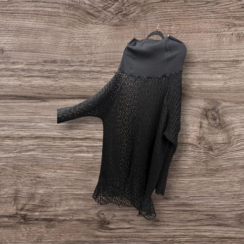 May include: A black, open-weave top with long sleeves and a high neckline. The top is hanging on a black hanger against a wood-grain background. The fabric has a textured, net-like pattern.