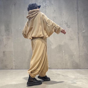 Op de afbeelding: Een beige tweedelige set bestaande uit een oversized hoodie met verlaagde schouders en een losvallende broek met wijde pijpen. De outfit wordt gecombineerd met zwarte plateauschoenen met een dikke, getextureerde zool en veters.