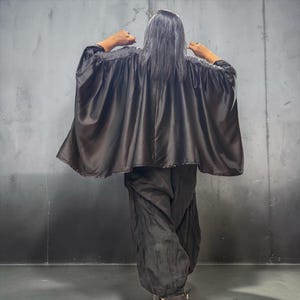 Puede incluir: Una capa negra con acabado satinado, mangas anchas y fluidas y escote fruncido, combinada con pantalones negros holgados. La prenda presenta una textura suave y reflectante con una silueta voluminosa que cae hasta la zona de la espalda.