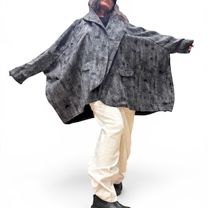 May include: A gray, oversized coat with a textured pattern and two front pockets. The coat has a unique, asymmetrical design and is paired with cream-colored pants and black boots. A black cap is worn on the head.