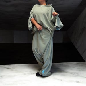 Op de afbeelding: Een lichtgroene oversized sweatshirt en bijpassende harembroek. De sweatshirt heeft wijde, uitlopende mouwen. De broek heeft een lage kruis en is bij de enkels gerimpeld. Een klein blauw ontwerp is zichtbaar op het linker broekspijp. De outfit is gemaakt van een zachte stof.