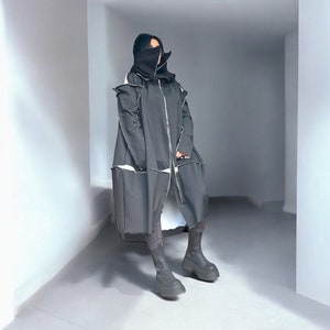 May include: A black hooded coat with white stitching and a zipper closure. The coat is worn by a person with a black face covering. The person is standing in a white hallway.