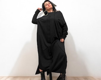 May include: A black long-sleeved maxi dress with a loose fit and a low neckline. The dress has a belt and a slit on the side. The model is wearing black boots.