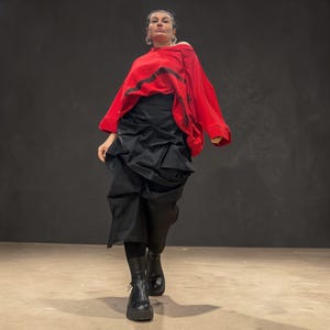 Könnte beinhalten: Eine Person trägt einen roten Kapuzenpullover mit schwarzen Akzenten, einen schwarzen Rock mit einem einzigartigen drapierten Design und schwarze Stiefel mit dicken Sohlen. Das Outfit befindet sich vor einem dunkelgrauen Hintergrund.