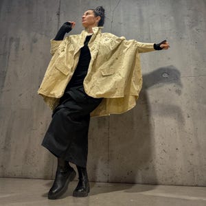 Puede incluir: Chaqueta beige texturizada con un diseño único y fluido, con bolsillos grandes. La chaqueta se combina con un atuendo negro, que incluye una falda larga y botas. La modelo está de pie frente a una pared de hormigón gris.