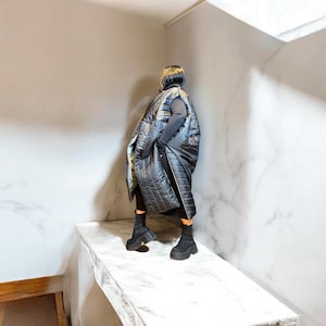 Könnte beinhalten: Eine schwarze Steppweste und schwarze Stiefel. Die lange Weste hat ein Steppmuster und einen Reißverschluss. Die Stiefel haben Plateausohlen. Das Outfit wird in einem Raum mit weißen Marmorwänden getragen.