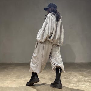 Puede incluir: Una sudadera con capucha gris claro de gran tamaño y pantalones de chándal anchos a juego. El conjunto se combina con botas de plataforma negras y una gorra de béisbol negra. Los pantalones tienen una raya blanca. El conjunto es informal y cómodo.