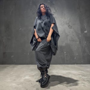 Könnte beinhalten: Ein dunkelgraues, übergroßes Kurzarm-Oberteil mit dezentem geometrischem Muster, kombiniert mit einer passenden, locker sitzenden dunkelgrauen Hose. Das Outfit wird durch schwarze Schnür-Plateaustiefel mit seitlichen Cut-outs ergänzt.