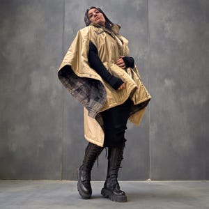 Peut inclure: Une veste cape à capuche beige avec une doublure à carreaux, portée sur une tenue noire. La veste a une fermeture éclair et des boutons. La personne porte des bottes noires à lacets et à plateforme. Le fond est un mur gris uni.