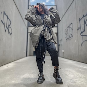 May include: An olive green jacket with a drawstring waist and long sleeves, paired with black trousers and black lace-up platform boots. The jacket features a gathered front closure with black ties, while the trousers have a relaxed fit.