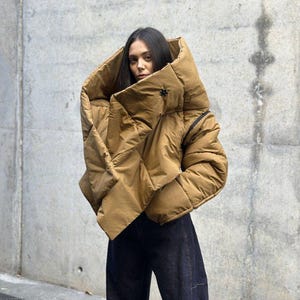 Puede incluir: Chaqueta acolchada color canela con cuello alto y cierre de botón. La chaqueta se lleva con pantalones vaqueros azul oscuro. La modelo está de pie frente a una pared de hormigón.
