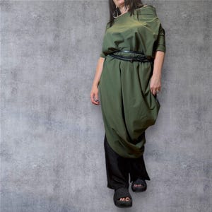 May include: An olive green dress with a black belt and black trousers. The dress features a unique draped design and short sleeves. The model is wearing black sandals. The background is a textured grey wall.