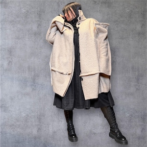 Può includere: Un cappotto beige, strutturato con un design a strati unico. Il cappotto presenta ampi revers e chiusure con bottoni. La persona indossa un vestito nero e stivali neri al ginocchio. Lo sfondo è grigio.