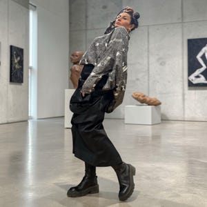 Op de afbeelding: Een persoon draagt een grijs en wit patroon trui, zwarte rok en zwarte laarzen. De persoon poseert in een moderne kunstgalerij met sculpturen en schilderijen op de achtergrond.