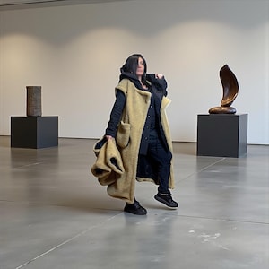 Puede incluir: Una persona lleva un chaleco largo con capucha beige con bolsillos grandes, sobre un atuendo negro. La persona está en una galería con esculturas sobre pedestales negros. El suelo es gris.