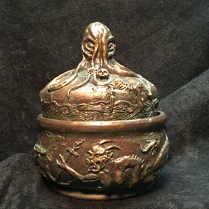 May include: A bronze-colored ceramic trinket box with a lid featuring a sculpted tentacled creature. The box is decorated with a relief design of a demon and other mythical creatures.
