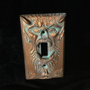 Puede incluir: Una placa de interruptor de luz de color bronce con un diseño detallado de rostro demoníaco. La cara tiene la boca abierta y el diseño presenta detalles turquesas. La placa del interruptor es rectangular con orificios para tornillos en la parte inferior.