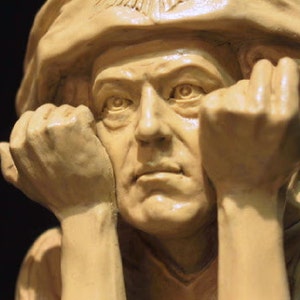 May include: A light beige sculpture of a man in a hat, hands supporting his face. The sculpture has detailed facial features and a thoughtful expression. The background is dark, highlighting the sculpture's form.