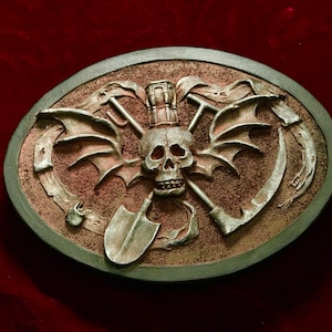 Puede incluir: Una hebilla de cinturón de metal plateado con una calavera, alas y palas cruzadas en relieve. El fondo es de color marrón texturizado.