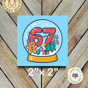 May include: A square magnet with a blue background features a snow globe design with the number 67 in red, a gingerbread person, and Christmas decorations. The magnet measures 2" x 2".
