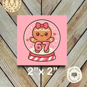 May include: A pink square magnet featuring a gingerbread person inside a snow globe. The gingerbread person has a pink bow and the number 67. The magnet measures 2 inches by 2 inches.