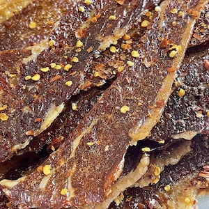 May include: Close-up of several pieces of beef jerky. The jerky is a dark brown color with visible marbling and is sprinkled with red pepper flakes and yellow seeds. The edges of the jerky appear slightly lighter in color and crispy.