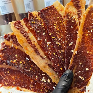 May include: Several thin, elongated slices of dark brown, cured meat arranged in a fan shape. The meat features a visible grain, a glossy surface, and is seasoned with scattered red chili flakes and small white pieces of dried garlic.