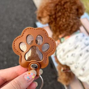 May include: A brown leather paw print keyring with a silver ring. The keyring has a stitched border and clear inserts in the paw pads. The background shows a brown dog.