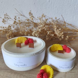 Puede incluir: Dos velas de hormigón blanco con adornos amarillos y rojos. Las velas tienen una mecha de madera y están etiquetadas "Talay candles" en una banda blanca. Una vela es más grande que la otra.
