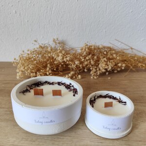 Puede incluir: Dos velas de hormigón blanco con mechas de madera y acentos florales secos. La vela más grande tiene forma ovalada y la más pequeña es redonda. Ambas velas tienen una etiqueta que dice "Talay candles".