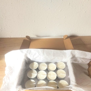 Puede incluir: Una caja llena de velas de té blancas, un total de 12, en una caja de cartón marrón con papel de seda blanco. La caja tiene el texto "Talay candles" impreso en la parte inferior.
