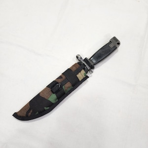 以下が含まれることがあります： 黒と緑の迷彩柄のナイフシースに、黒いナイフが入っています。