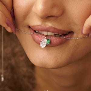 May include: A delicate silver chain necklace featuring a small oval pendant with an engraved design and a tiny, vibrant green gemstone accent. The necklace is held near the mouth, showcasing the fine metal link construction and the pendant detail.