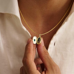 Puede incluir: Un collar con un colgante de medallón dorado. El colgante redondo está abierto, revelando una pequeña imagen y una piedra verde. El collar se lleva sobre una camisa blanca.