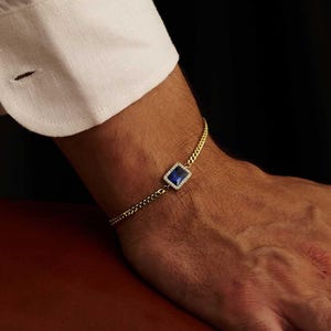 Peut inclure: Un bracelet en or avec une pierre précieuse carrée bleu foncé. Le bracelet est porté sur un poignet, à côté d'une manche de chemise blanche. Le fond est sombre.