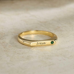 Peut inclure: Une bague dorée dotée d'une section plate et horizontale gravée du prénom Freya en écriture cursive, accompagnée d'une petite pierre précieuse ronde de couleur vert foncé incrustée dans le métal à côté de l'inscription.