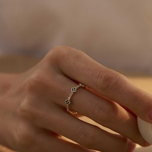 May include: A gold ring with black floral detailing adorns a hand holding a white teacup. The ring showcases small, repeating black flower shapes. The background has a neutral, warm tone, with a glimpse of a white shirt.