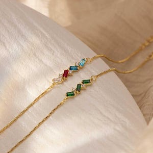 May include: Two delicate gold chain bracelets. One features a row of colorful rectangular gemstones in red, blue, and clear. The other has green gemstones. The bracelets are displayed on a soft, white fabric.