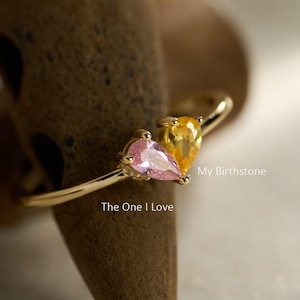 May include: A gold ring featuring two pear-cut gemstones, one pink and one yellow, set side-by-side. The text "My Birthstone" and "The One I Love" is printed near the stones, with "Handmade By H Jewellery Store" at the bottom.