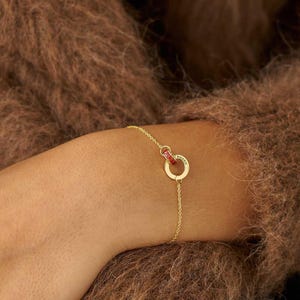 Könnte beinhalten: Ein zartes Goldkettenarmband mit einem kreisförmigen Anhänger, an dem ein kleiner roter Akzentring befestigt ist. Die Goldkette ist dünn und minimalistisch und mit dem kreisförmigen Anhänger verbunden, der eine glatte, polierte Oberfläche aufweist.