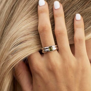May include: A silver ring featuring a rectangular, emerald-cut green gemstone set in a smooth, polished metal band. The ring has a double-band appearance, with the stone centered on the top section, showcasing a clean, metallic finish.