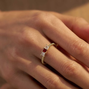 May include: A gold ring featuring a beaded band and three square-cut stones, with a central red stone flanked by two clear white stones. The ring is positioned on a finger, and the text Handmade By H Jewellery store is visible at the bottom.