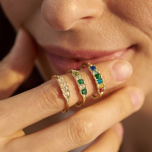 May include: Three gold rings, each set with a row of coloured gemstones. The rings feature clear, green, and rainbow-coloured stones. The rings are displayed on a finger, showcasing the jewellery's design and colour variations.
