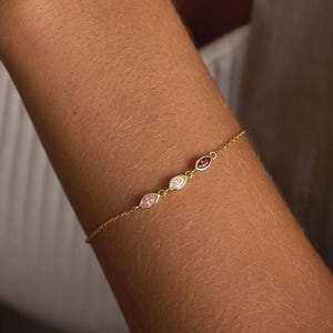 Peut inclure: Un bracelet en or fin orné de trois pierres précieuses serties en serti clos, de couleurs rose, blanche et rouge. Le bracelet est présenté sur un bras, avec les pierres espacées le long de la chaîne. Il s'agit d'un bijou.