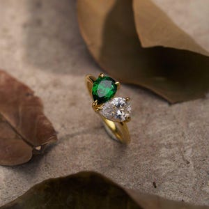 May include: A gold ring featuring two pear-cut stones, one vibrant green and one clear white, set in a bypass style. The band is polished gold metal. The text "Handmade By H Jewellery store" is printed in small white letters at the bottom.