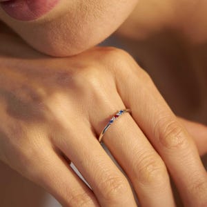 May include: A thin silver-toned metal ring featuring three small, round, prong-set gemstones arranged in a row. The stones consist of a blue gem on the left, a red gem in the centre, and a blue gem on the right, set on a smooth, slender band.
