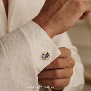 Puede incluir: Un gemelo de tono dorado con una piedra preciosa azul rectangular engastada en un marco ovalado, sujeto en el puño de una camisa blanca de manga larga. El diseño incluye una piedra azul central rodeada por un borde de metal dorado pulido.